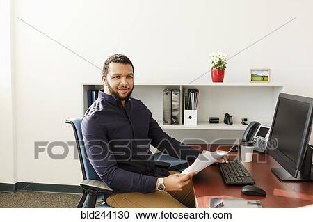 Mixed Race man reading paperwork near computer in office View Large Photo Image Stock Image - Mixed Race man reading paperwork near computer in office. Fotosearch