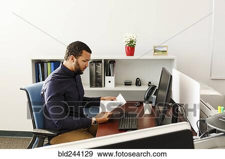 Stock Photo - Mixed Race man reading paperwork near computer in office. Fotosearch