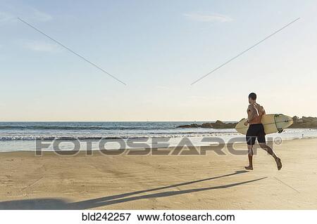 Stock Photo - Mixed race man running with surfboard on beach. Fotosearch