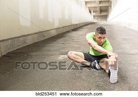 Stock Image - Mixed Race man sitting on ground stretching leg. Fotosearch