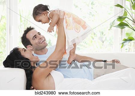 Stock Image - Mixed Race mother and father playing on sofa with baby daughter. Fotosearch