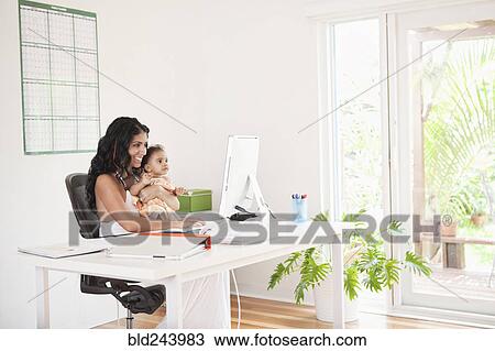 Stock Image - Mixed Race mother using computer and holding baby daughter. Fotosearch