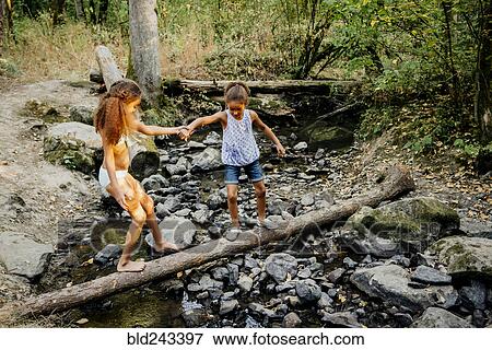 Stock Photo - Mixed Race sisters holding hands on log over forest stream. Fotosearch