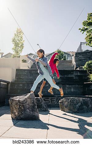 Stock Photograph - Mixed Race sisters jumping on rocks. Fotosearch