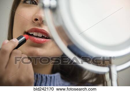 Stock Image - Mixed Race woman applying lipstick in mirror. Fotosearch