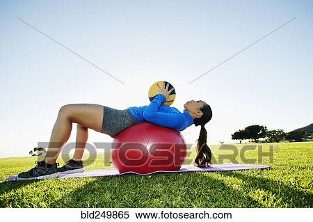 Stock Photography - Mixed Race woman exercising with fitness ball and heavy ball in sunny field. Fotosearch