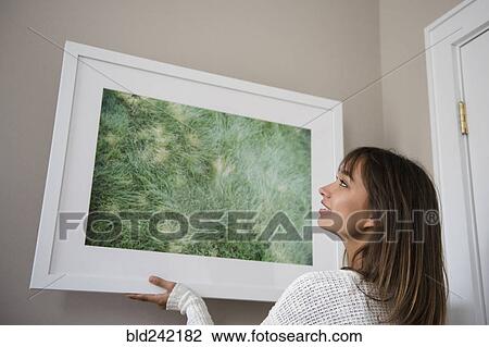 Stock Image - Mixed Race woman hanging picture frame on wall. Fotosearch