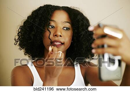 Stock Photograph - Mixed Race woman holding cell phone and applying lipstick. Fotosearch