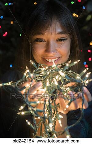Mixed Race woman holding string lights near Christmas tree View Large Photo Image Picture - Mixed Race woman holding string lights near Christmas tree. Fotosearch