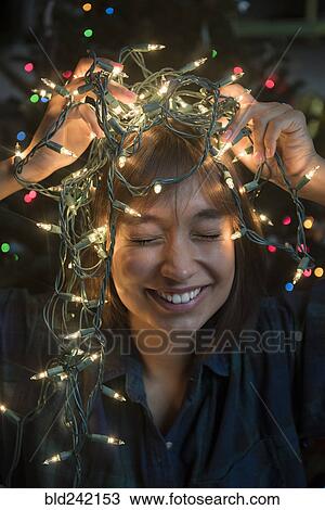 Stock Image - Mixed Race woman holding string lights on head near Christmas tree. Fotosearch