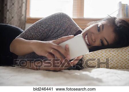 Stock Image - Mixed Race woman laying on bed texting on cell phone. Fotosearch
