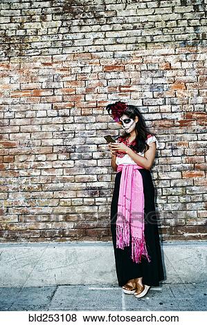 Stock Photo - Mixed Race woman on sidewalk wearing skull face paint texting on cell phone. Fotosearch