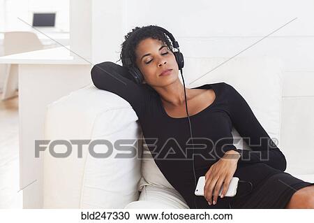 Stock Image - Mixed Race woman on sofa listening to cell phone with headphones. Fotosearch