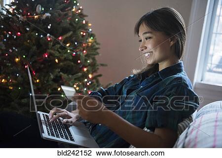 Stock Image - Mixed Race woman online shopping with laptop near Christmas tree. Fotosearch