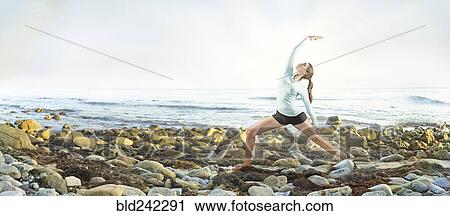 Stock Image - Mixed race woman practicing yoga on rocky beach. Fotosearch