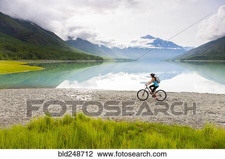 Stock Image - Mixed Race woman riding bicycle near lake. Fotosearch