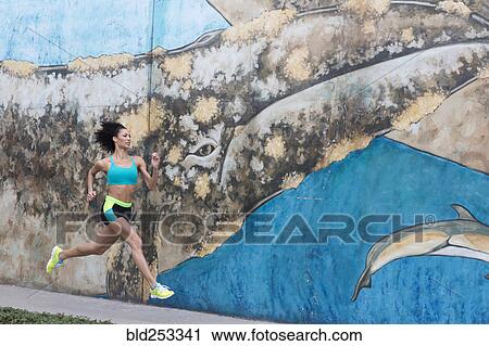 Stock Image - Mixed Race woman running on sidewalk near mural. Fotosearch
