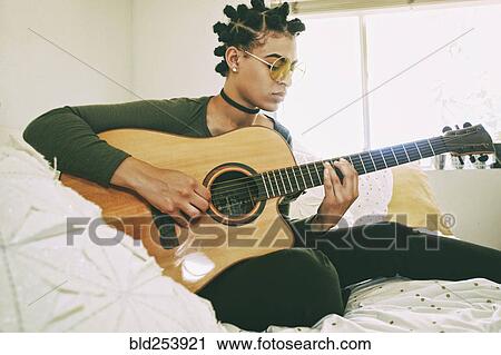 Stock Image - Mixed Race woman sitting on bed playing guitar. Fotosearch