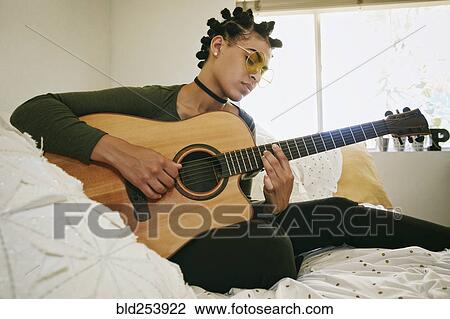 Stock Image - Mixed Race woman sitting on bed playing guitar. Fotosearch