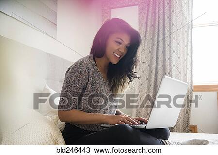 Stock Image - Mixed Race woman sitting on bed using laptop. Fotosearch