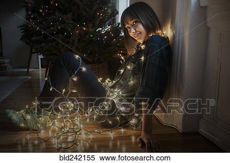 Stock Photography - Mixed Race woman sitting on floor wrapped in string lights near Christmas tree. Fotosearch