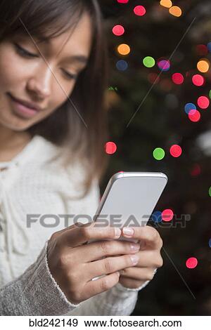Stock Photo - Mixed Race woman texting on cell phone near Christmas tree. Fotosearch