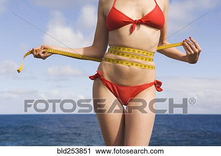 Stock Image - Mixed Race woman wearing bikini measuring waist near ocean. Fotosearch