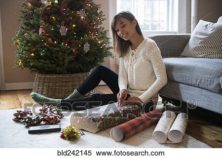 Stock Photography - Mixed Race woman wrapping gift on floor near Christmas tree. Fotosearch