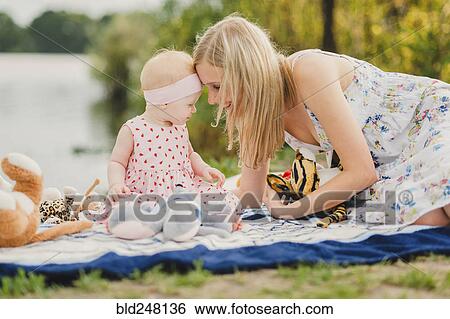 Stock Photograph - Mother and daughter playing with stuffed animals on blanket near lake. Fotosearch