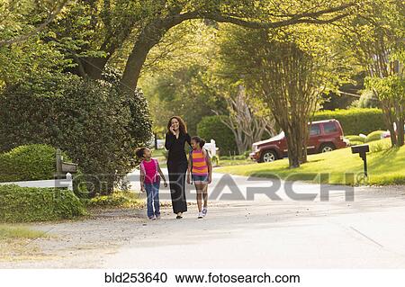 Stock Image - Mother walking in street with daughters wearing backpacks. Fotosearch