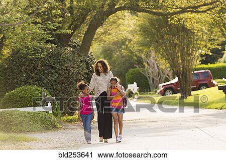 Stock Image - Mother walking in street with daughters wearing backpacks. Fotosearch
