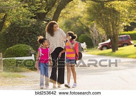 Stock Image - Mother walking in street with daughters wearing backpacks. Fotosearch