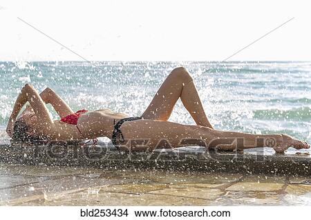 Picture - Ocean waves splashing on Caucasian woman wearing bikini. Fotosearch