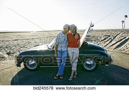 Stock Photo - Older Caucasian couple leaning on convertible car with surfboard on beach. Fotosearch