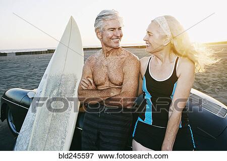 Stock Photo - Older Caucasian couple leaning on convertible car with surfboards. Fotosearch