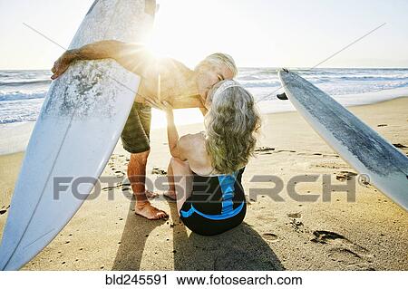 Stock Image - Older Caucasian couple on beach with surfboards kissing. Fotosearch