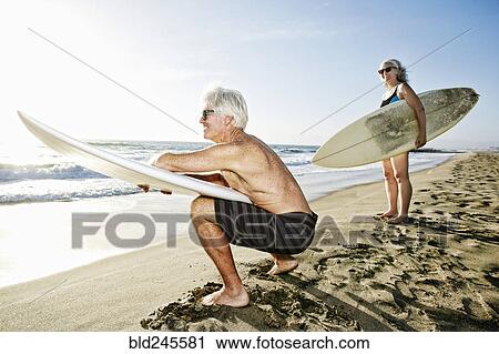 Stock Image - Older Caucasian couple standing on beach holding surfboards. Fotosearch