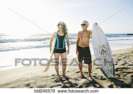 Stock Photo - Older Caucasian couple standing on beach holding surfboards. Fotosearch