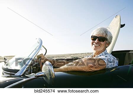 Picture - Older Caucasian man in convertible car with surfboard on beach. Fotosearch