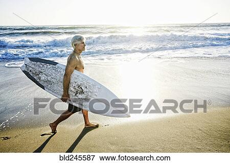 Stock Photo - Older Caucasian man walking on beach carrying surfboard. Fotosearch