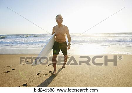 Stock Photo - Older Caucasian man walking on beach carrying surfboard. Fotosearch