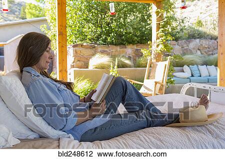 Stock Image - Older Caucasian woman laying on bed reading book in garden. Fotosearch