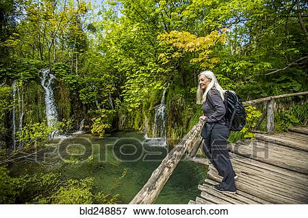 Stock Photo - Older Caucasian woman on wooden bridge admiring waterfalls. Fotosearch