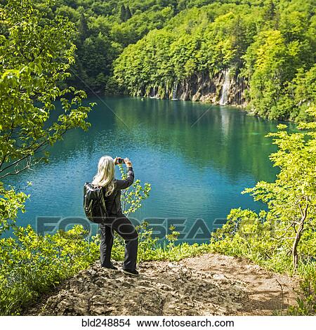 Picture - Older Caucasian woman photographing waterfall with cell phone. Fotosearch
