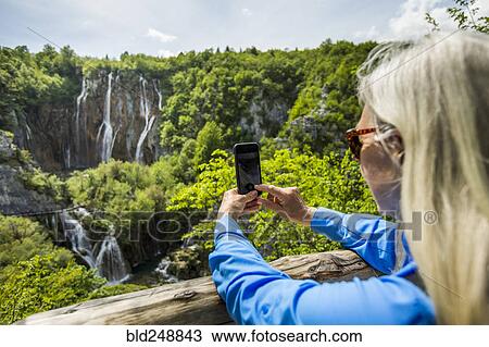 Stock Image - Older Caucasian woman photographing waterfalls with cell phone. Fotosearch