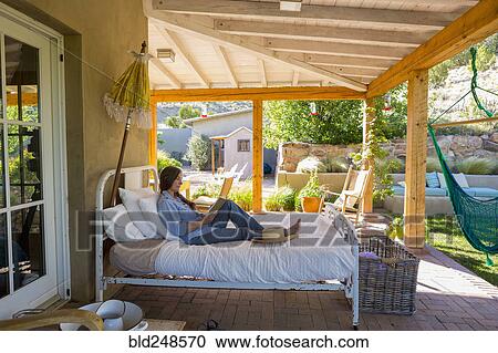 Stock Image - Older Caucasian woman reading books on bed on patio. Fotosearch