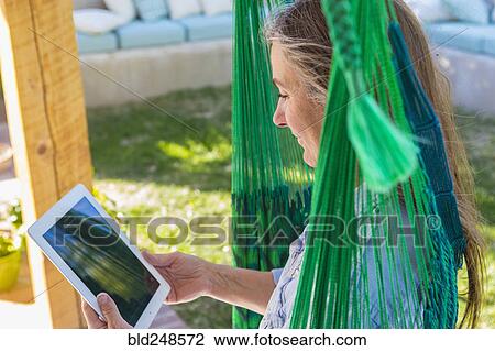 Stock Image - Older Caucasian woman reading digital tablet in green hammock. Fotosearch