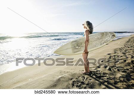 Stock Image - Older Caucasian woman standing on beach holding surfboard. Fotosearch