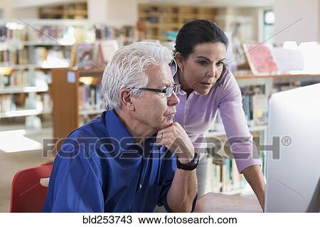 Stock Image - Older Hispanic woman helping man using computer in library. Fotosearch
