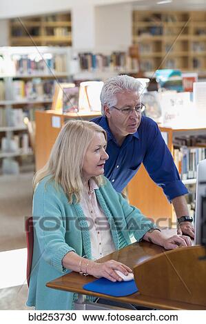 Stock Image - Older man helping woman using computer in library. Fotosearch